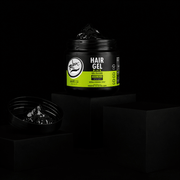 Black jar of Hair Gel (Black) styling gel placed on matte black blocks against a black background, with the lid off showing glossy gel texture