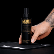 Hand holding a STMNT Definition Spray Bottle on a Black mat on a black background Primos Barber Supply