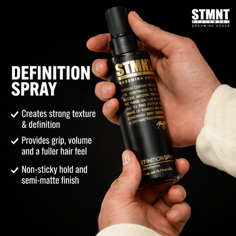 Two Hands holding a STMNT Definition Spray bottle with product details on a black background Primos Barber Supply
