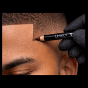 Person applying a brown L3VEL3 pencil to their skin wearing black gloves on a black background.