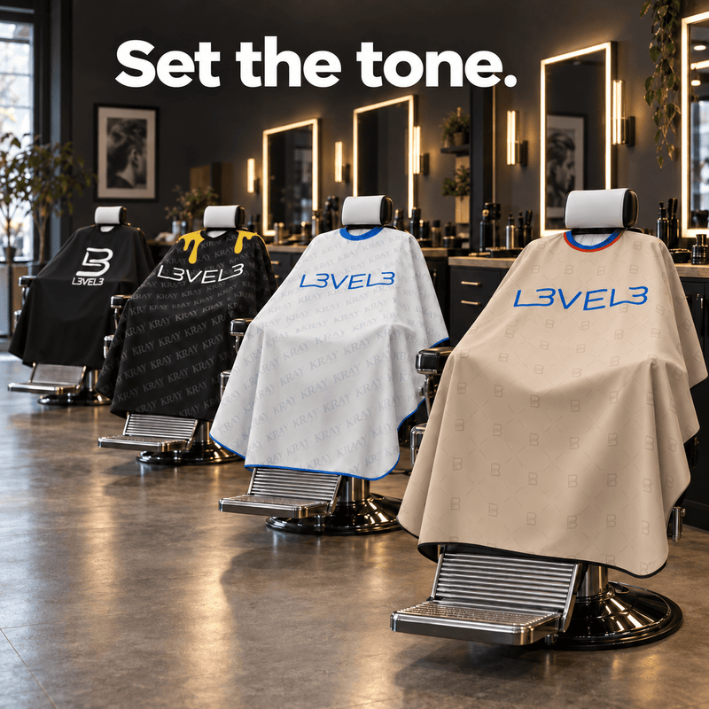 Four L3VEL3 rubber neck cutting capes in black, black drip, white, and beige draped over barber chairs in a shop
